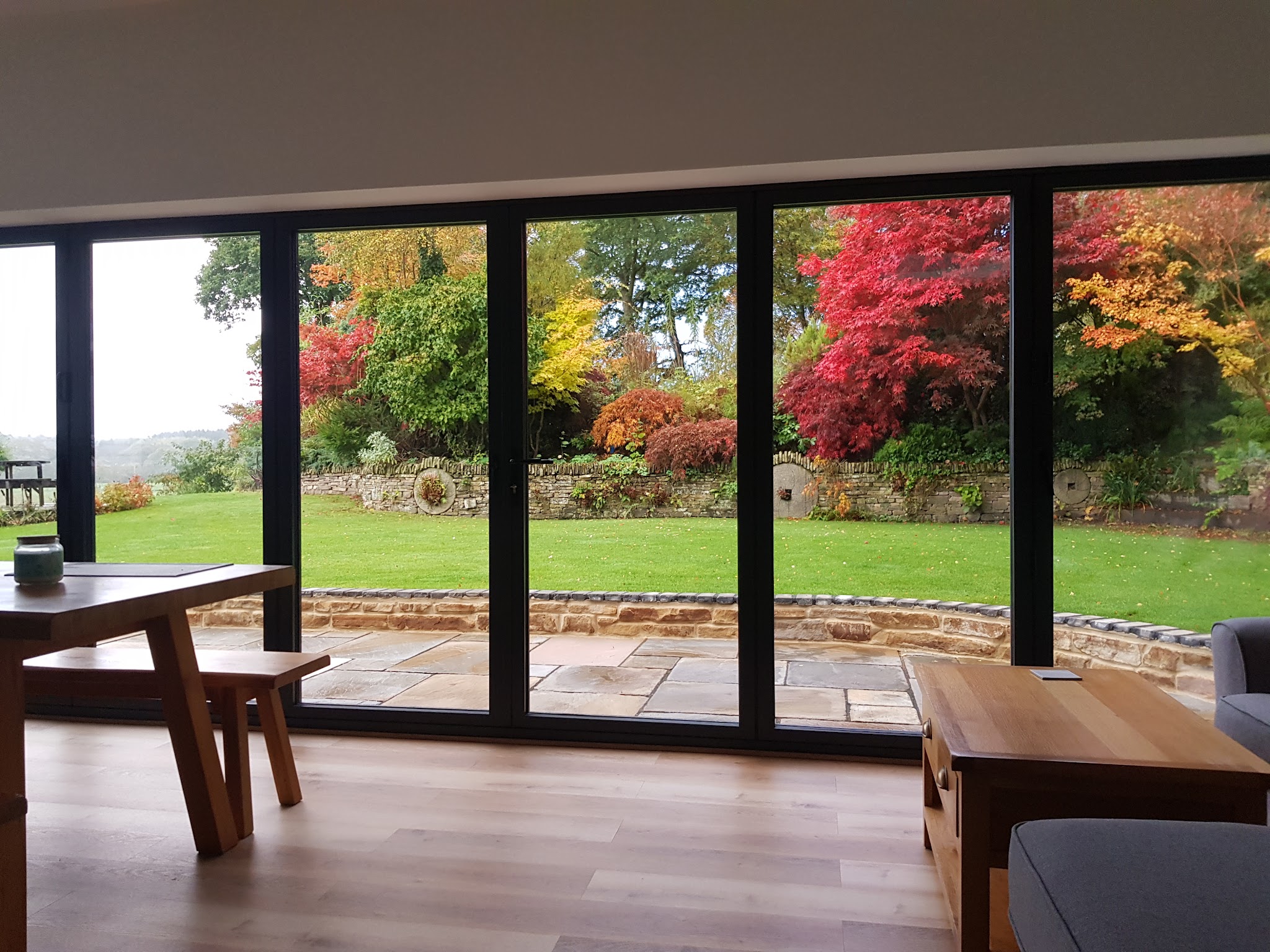 View from a dining room through anthracite folding patio doors onto a lawn and autumn trees